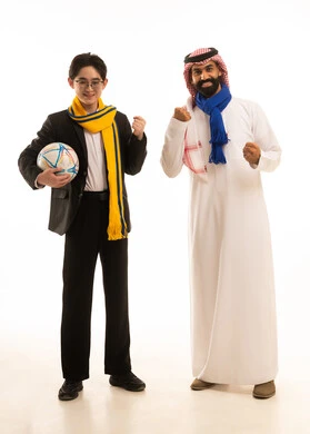 بورتريه لشاب اسيوى يرتدي البدلة الرسمية و وشاح فريق النصريحمل بيديه كورة القدم  يقف بجانب رجل عربي خليجي سعودى يرتدي الثوب التقليدى والشماغ و وشاح فريق الهلال يرفعان يديهما لتشجيع فريق كرة القدم، صورة الجسم بالطول الكامل، خلفية بيضاء