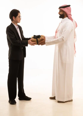 مفهوم الشكر والامتنان، التعبير عن الحب والتقدير والاحترام، بورتريه لرجل عربي خليجي سعودى يرتدي الثوب التقليدى و الشماغ يقوم بتقديم هدية لصديق اسيوى يرتدي البدلة الرسمية، ايماءات السعادة والسرور، الجسم بالطول الكامل، خلفية بيضاء