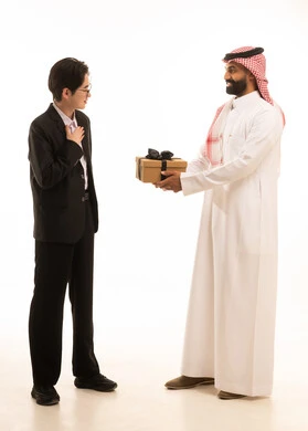 بورتريه لرجل عربي خليجي سعودى يرتدي الثوب التقليدى و الشماغ يقوم بتقديم هدية لصديق اسيوى يرتدي البدلة الرسمية، يماءات السعادة والدهشة والسرور، مشاعر الحب بين الاصدقاء، الجسم بالطول الكامل، خلفية بيضا