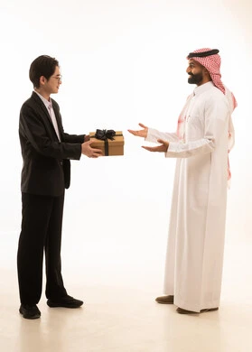 تقديم الهدايا التذكارية, التعبير عن المحبة والألفة، بورتريه لشاب اسيوى يرتدي البدلة الرسمية يقوم بتقديم هدية لرجل عربي خليجي سعودى يرتدي الثوب التقليدى و الشماغ ، ايماءات السعادة والدهشة والسرور، الجسم بالطول الكامل، خلفية بيضاء