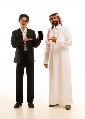 A black screen displayed on a mobile phone, a portrait of a Saudi Gulf Arab man wearing traditional attire standing next to an Asian young man in a formal suit pointing at the phone, using electronic programs and applications, full-body shot, white background.