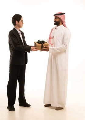 مشاعر الحب بين الاصدقاء، بورتريه لرجل عربي خليجي سعودى يرتدي الثوب التقليدى و الشماغ يقوم بتقديم هدية لصديق اسيوى يرتدي البدلة الرسمية، يماءات السعادة والدهشة والسرور، الجسم بالطول الكامل، خلفية بيضاء