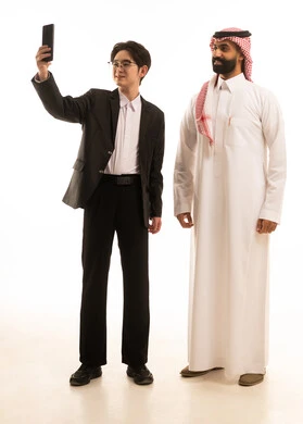 Taking a commemorative selfie via mobile, a portrait of a Saudi Arabian Gulf man wearing traditional attire standing next to a young Asian man in formal suit holding a mobile phone, using electronic programs and applications, full body, white background.