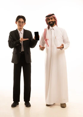 التسويق لمنتج ما، عرض شاشة سوداء عبرالجوال، بورتريه لرجل عربي خليجي سعودى يرتدي الثوب التقليدى يقف بجانب شاب اسيوى يرتدي البدلة الرسمية، استخدام البرامج والتطبيقات الالكترونية، الجسم بالطول الكامل، خلفية بيضاء