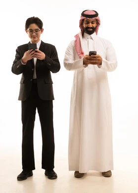 بورتريه لرجل عربي خليجي سعودى يرتدي الثوب التقليدىر شاب اسيوى يرتدي البدلة الرسمية يمسك الهاتف و يقف بجانب شاب اسيوى يرتدي البدلة الرسمية يستخدم الجوال، إيماءات الاندماج والتركيز، الجسم بالطول الكامل، خلفية بيضاء