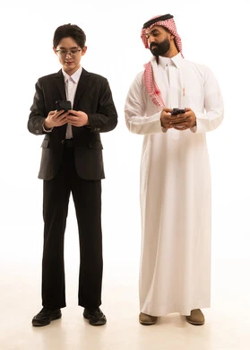 إيماءات الاندماج والتركيز، بورتريه لرجل عربي خليجي سعودى يرتدي الثوب التقليدىر شاب اسيوى يرتدي البدلة الرسمية يمسك الهاتف و يقف بجانب شاب اسيوى يرتدي البدلة الرسمية يستخدم الجوال، الجسم بالطول الكامل، خلفية بيضاء