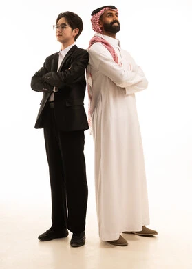 بورتريه لرجل عربي خليجي سعودى يرتدي الثوب التقليدى والشماغ يقف و ظهره الى ظهر شاب اسيوى يرتدي البدلة الرسمية ينظران الى شىء ما، الوقوف بأيدى متقاطعة، الجسم بالطول الكامل، خلفية بيضاء