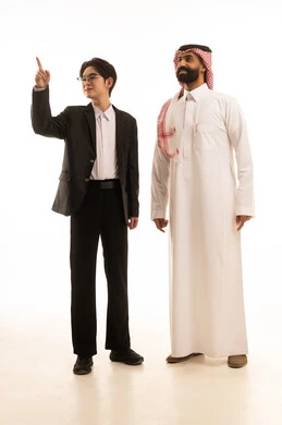 Pointing to something, a portrait of a young Asian man in a formal suit pointing with his hands to a certain place and looking at a Saudi Arabian Gulf Arab man wearing traditional attire and a ghutrah standing next to him, a gesture indicating this place, full-body image, white background.