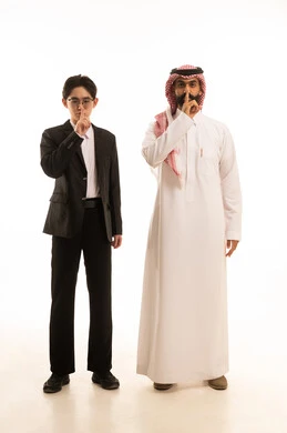 Looking at the camera with expressions requesting silence, a portrait of a Saudi Arabian Gulf man wearing traditional attire and a ghutrah standing next to a Chinese young man in a formal suit, both raising their fingers in gestures for silence, indicating to lower the volume, white background, full-body image, white background.