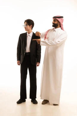 تعابير التحديد والتأكيد, الاشارة إلى شيء ما بإيماءات السرور ,بورتريه لرجل عربي خليجي سعودي مبتسم يرتدي الزي التقليدي والشماغ يشير بأصابع السبابة للاختيار و يضع يده على كتف شاب اسيوى يرتدى البدلة الرسمية لرؤية شىء ما، الجسم بالطول الكامل، خلفية بيضاء