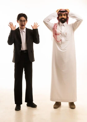 حركة وجه وفم تدل على الصدمة و الدهشة، بورتريه لرجل عربي خليجي سعودى يرتدي الثوب التقليدى و الشماغ يتحاور مع شاب اسيوى يرتدي البدلة الرسمية يرفعان أيديهم للأعلى بتعابيرالتفاجؤ لرؤية شيء ما, صورة الجسم بالطول الكامل، خلفية بيضاء .