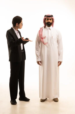 مفهوم التساؤل والحيرة ، بورتريه لشاب صينى يرتدي البدلة الرسمية يرفع كفيه و ينظر بتعجب لرجل عربي خليجي سعودى يرتدى الثوب التقليدى والشماغ يقف فى غموض و ثبات، صورة الجسم بالطول الكامل، خلفية بيضاء