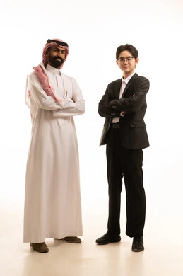 Standing with crossed arms, a portrait of a Saudi Arabian Gulf man wearing traditional attire and a shemagh stands next to a young Chinese man in a formal suit, both looking at the camera with expressions of joy, exchanging experience and knowledge between countries, hosting foreign businessmen in the Kingdom, white background.