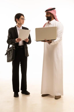 Hosting foreign businessmen in the Kingdom, using modern technology to accomplish work, signing a new work agreement, a portrait of a Saudi Gulf Arab man wearing traditional attire holding a laptop standing next to a Chinese young man wearing a formal suit and carrying a black bag with work reports, white background.