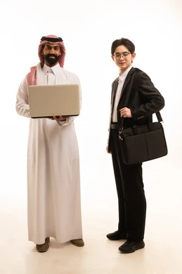 A portrait of a young Asian man wearing a formal suit standing next to a Saudi Arabian Gulf man dressed in traditional thobe and ghutrah, both working on a laptop, exchanging experience and knowledge between countries, with a white background.