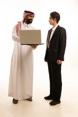 Exchange of experience and knowledge between countries, a portrait of a Saudi Arabian Gulf man wearing traditional attire and a shemagh standing next to an Asian young man in a formal suit, both working on a laptop, with a white background.