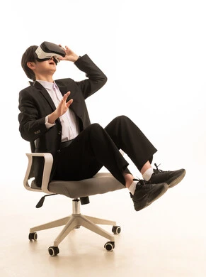 Using a modern and advanced technical device, the enjoyment and entertainment of watching through virtual reality glasses, a portrait of an Asian young man wearing a formal suit sitting on a chair looking through 3D glasses with expressions of immersion and excitement, white background.