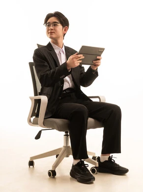 Looking at something, using a modern advanced technical device, a portrait of an Asian man wearing a formal suit sitting on a chair holding a tablet browsing the internet, remote work, fast and secure online shopping, white background.