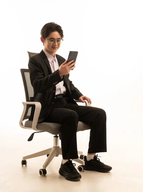 A portrait of an Asian man wearing a formal suit sitting on a comfortable chair holding a mobile phone with expressions of happiness, using modern technology, sending text and voice messages via mobile, white background.