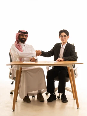 استضافة رجل اعمال اجنبى في السعودية، تبادل الخبرة والمعرفة بين البلاد, بورتريه لرجل عربي خليجي سعودى يرتدي الثوب التقليدى و الشماغ يجلس على طاولة الاجتماع يصافح رجل اسيوى يرتدي البدلة الرسمية، خلفية بيضاء