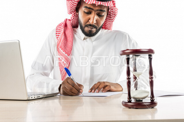 بورتريه لرجل أعمال سعودي خليجي في المكنب ، استخدام جهاز الحاسوب المحمول ، الساعة الرملية الزجاجية ( البنكام ) على الطاولة ، جهاز قياس مرور ونفاد الوقت ، الساعة الرملية الخشبية ، مفهوم إدارة الأعمال والوقت ، مهن ووظائف مكتبية ، إنجاز المهام بدقة وكفاءة