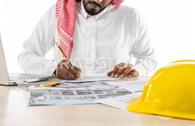 بورتريه  لمهندس خليجي سعودي داخل مكتب هندسي ، إستخدام جهاز الحاسوب المحمول للإنجاز وتتبع المهام ، خوذة الوقاية ، استخدام أدوات ومعدات هندسية  على مخطط تصميم هندسي ، مهن ووظائف هندسية ، خلفية بيضاء ، مفهوم الهندسة المعمارية ، مهندس معماري محترف