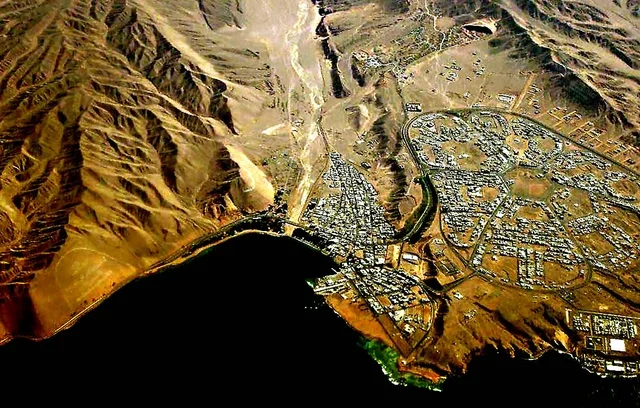 A coastal city surrounded by mountainous terrain, an aerial view of the city of Haql in the Tabuk region of Saudi Arabia, located on the Gulf of Aqaba, the extension of urban areas along the coast with the slope of the surrounding mountains, buildings and streets are spread out in organized geometric divisions.