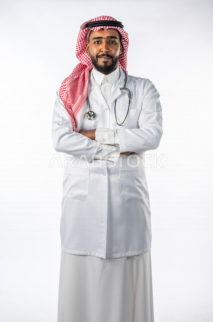 بورتريه لرجل عربي خليجي سعودي ، ينظر إلى الكاميرا بإيدي متقاطعة ، يرتدي المعطف الطبي الخاص بمهنة الطبيب ، مفهوم الطب والرعاية الصحية ، خلفية بيضاء ، مهن و وظائف طبية ، العمل في القطاع الصحي لتقديم الخدمات الصحية ، يقدم خدماته للمرضى بكل كفاءه