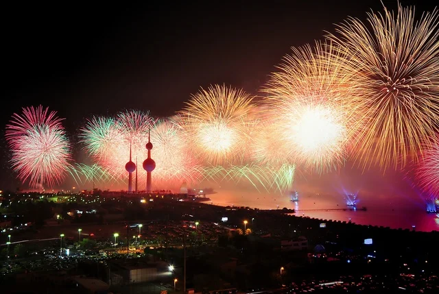 Fireworks and games above the sky of Kuwait, the architectural engineering art of the towers of Kuwait City, skyscrapers and facilities on the coast of the Arabian Gulf in the Ras Ajouzah area, urban development and progress in a modern and advanced style, green trees and plants, a destination for visitors from around the world.