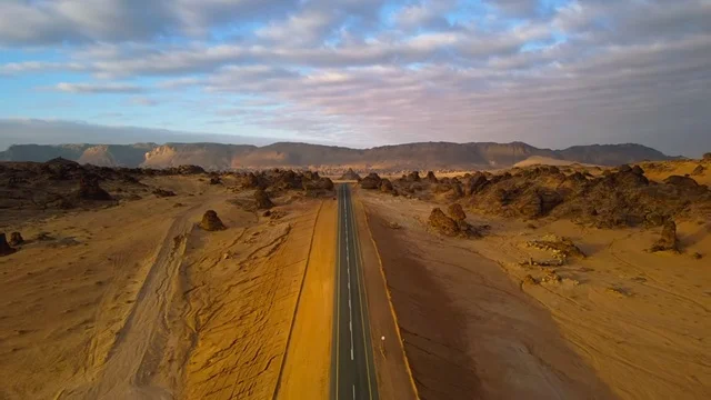 Aerial drone footage of a straight road in Madain Saleh in Al-Ula Province, Saudi Arabia, showcasing the desert nature, the heritage city of Al-Ula, the historical castles of Al-Ula, driving through the desert at sunset, and ancient historical archaeological landmarks.