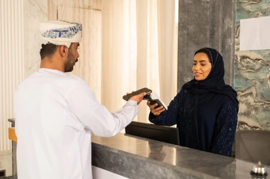 إجراءات الحجز والدخول لدى مكتب الاستقبال في الفندق، سياحه وفندقه وسفر، رجل عربي خليجي عماني يرتدي الدشداشة و العمامة يقوم بالدفع عند الكاشير بطريقة الكترونية بإستخدام الجوال، إستعلامات وإستفسارات فى خدمة العملاء