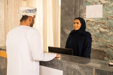An Arab Gulf Omani man wearing a dishdasha and a turban is inquiring at customer service, completing the booking procedures and checking into the hotel, with expressions of happiness and joy, the concept of tourism, hospitality, and travel.