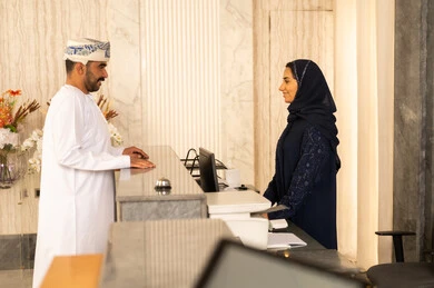 Expressions of happiness and joy, completing the booking procedures and checking into the hotel, the concept of tourism, hospitality, and travel, an Arab Gulf Omani man wearing a dishdasha and a turban inquiring at customer service.