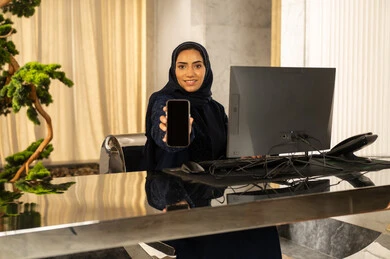 عرض شاشة سوداء فارغة، جودة تطبيقات وخدمات الهاتف المحمول، استخدام التقنيات الحديثة، امراه عربيه خليجيه سعودية محجبة ترتدي العباءة تجلس امام الحاسوب , النظر الى الكاميرا بايماءات السعادة و السرور، استقبال الضيوف وإتمام الحجز