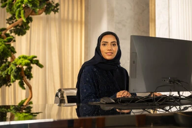 انجاز مهام العمل، امراه عربيه خليجيه سعودية محجبة ترتدي العباءة  تعمل على جهاز الحاسوب، النظر الى الكاميرا بتعابير السرور، الإجابة عن الاستعلامات والاستفسارات، جذب واستقطاب المزيد من العملاء، العمل على زيادة مستوى رضا العميل