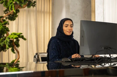 Typing on a keyboard, a Saudi Arabian Gulf woman wearing a hijab and an abaya is working on a computer, showing gestures of engagement and focus, answering inquiries and questions, attracting and drawing in more customers, and working to increase customer satisfaction levels.