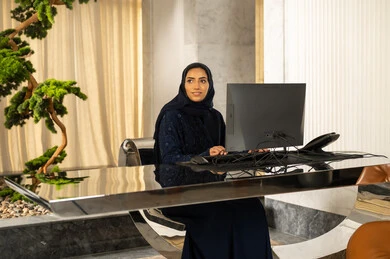 Looking at something, answering inquiries and questions, attracting and drawing more customers, working to increase customer satisfaction, an Arab Gulf Saudi woman wearing a hijab and an abaya working on a computer.