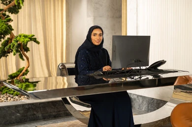 Looking at the camera with expressions of joy, answering inquiries and questions, attracting and drawing in more customers, working to increase customer satisfaction, an Arab Gulf Saudi woman wearing a hijab and an abaya working on a computer.