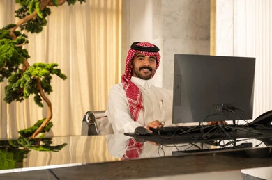 النظر الى الكاميرا بتعابير السرور، جذب واستقطاب المزيد من العملاء، العمل على زيادة مستوى رضا العميل، الإجابة عن الاستعلامات والاستفسارات, رجل عربي خليجي سعودي يرتدي الثوب الابيض و الشماغ يعمل على جهاز الحاسوب