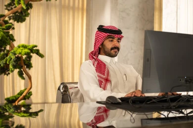 Attracting and recruiting more customers, working to increase customer satisfaction, answering inquiries and questions, an Arab Gulf Saudi man wearing a white thobe and a shemagh working on a computer.