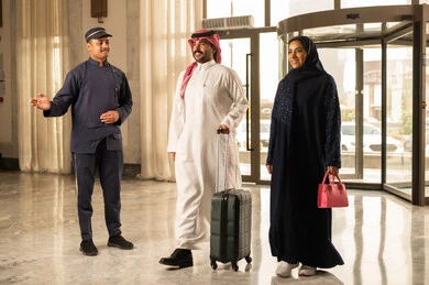 رحلة عائلية ترفيهية، الترحيب بالضيوف والبشاشة، ايماءات السعادة و الحماس، رجل عربي خليجي سعودي يرتدي الثوب الابيض و الشماغ يمسك بيديه حقيبة السفر يدخل الى الفندق برفقه زوجته ، قضاء أجمل الأوقات في أفضل الفنادق الخليجية