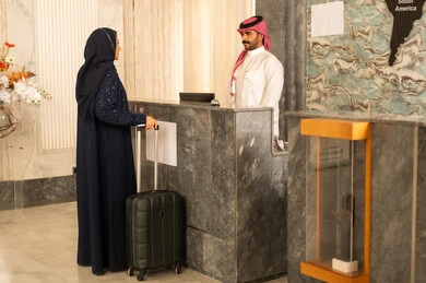 Checking in at the hotel reception, the concept of tourism, hospitality, and travel, an Arab Gulf woman from Saudi Arabia wearing a hijab and an abaya holding a travel bag in her hands is inquiring at customer service.