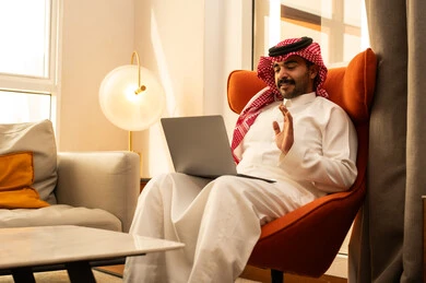 Raising the palm of the hand with expressions of greeting and peace, making a video call, the concept of remote work, the evolution of technology and technical devices, using a laptop to accomplish tasks and work, an Arab Gulf Saudi man in a white thobe and ghutrah sitting in a hotel next to a suitcase using a laptop.