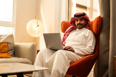 Looking at the camera with expressions of joy, the concept of remote work, the evolution of technology and technical devices, using a laptop to accomplish tasks and work, an Arab Gulf Saudi man in a white thobe and ghutrah sitting in a hotel next to a travel bag using a laptop.