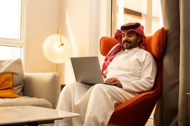 The concept of remote work, the evolution of technology and technical devices, using a laptop to accomplish tasks and work, an Arab Gulf Saudi man in a white thobe and ghutrah sitting in a hotel next to a travel bag using a laptop.