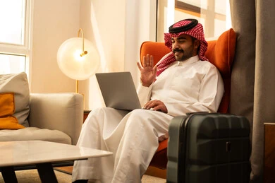 اجراء مكالمة فيديو، رفع كف اليد بتعابير التحية و السلام، مفهوم العمل عن بعد، تطور التكنولوجيا والأجهزة التقنية، استخدام اللابتوب لانجاز المهام والعمل، رجل عربي خليجي سعودي الثوب الابيض و الشماغ يجلس في الفندق بجانب حقيبة السفر يستخدم الحاسوب المحمول