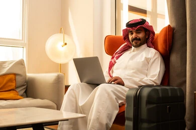 The development of technology and technical devices, the use of laptops to accomplish tasks and work, an Arab Gulf Saudi man in a white thobe and shemagh sitting in a hotel next to a travel bag using a laptop, the concept of remote work.