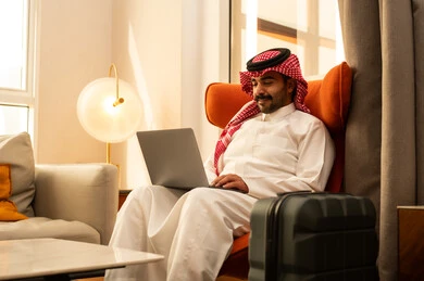 Using a laptop to accomplish tasks and work, an Arab Gulf Saudi man in a white thobe and shemagh sits in a hotel next to a travel bag using a laptop, the evolution of technology and technical devices, the concept of remote work.
