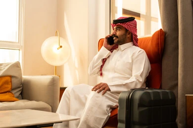 اجراء مكالمة هاتفية عبر الجوال، الدردشة مع الاهل و الاصدقاء، تطور التكنولوجيا والأجهزة التقنية، رجل عربي خليجي سعودي الثوب الابيض و الشماغ يجلس في الفندق بجانب حقيبة السفر يستخدم الهاتف المحمول