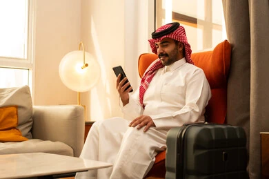 Taking commemorative selfies, live streaming with family and friends, using a mobile phone, integrating modern technologies into daily life, browsing social media, an Arab Gulf Saudi man in a white thobe and ghutrah sitting in a hotel next to a suitcase holding a mobile phone in his hand.